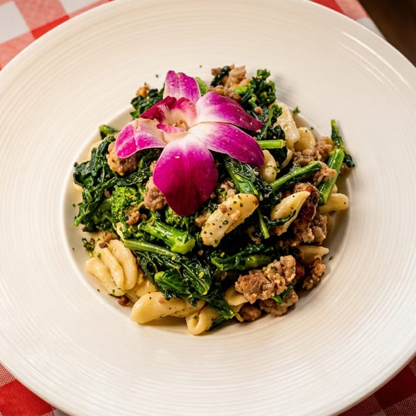 A white plate holds pasta with broccoli rabe, sausage, and a pink orchid on a checkered tablecloth.