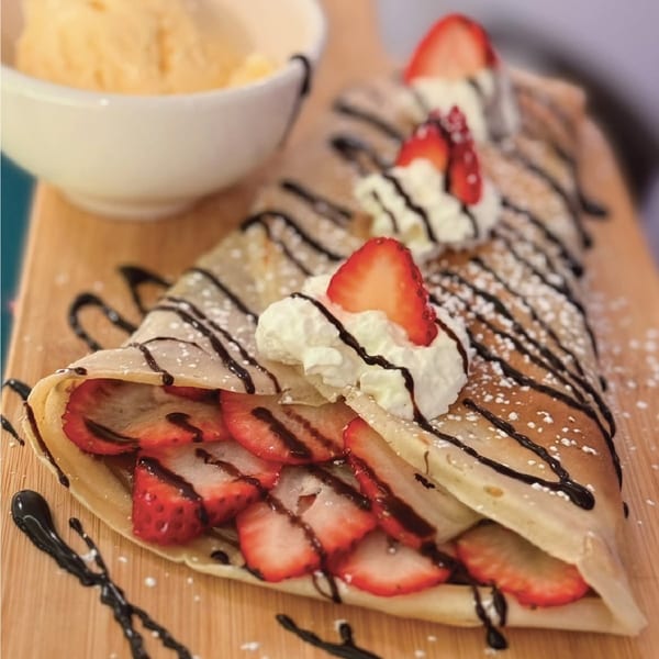 Strawberry crepe with whipped cream, chocolate drizzle, powdered sugar, and a side of ice cream on a wooden board.