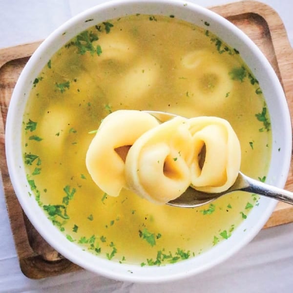 A spoon lifts tortellini from a white bowl of clear broth soup with parsley.