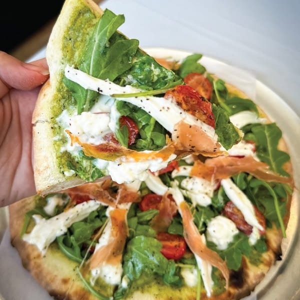 Hand lifting a slice of pesto pizza with arugula, tomatoes, mozzarella, and prosciutto.